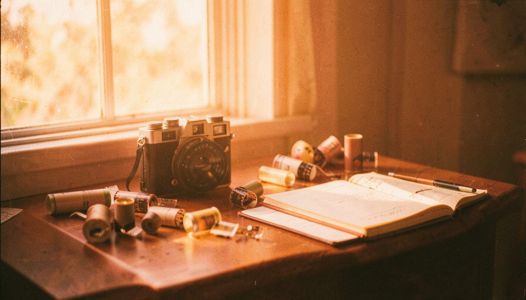 Warm orange-toned analog film photograph of a wooden desk beside a sunlit window, with a vintage camera, scattered film rolls and a notebook, soft golden daylight and visible grain