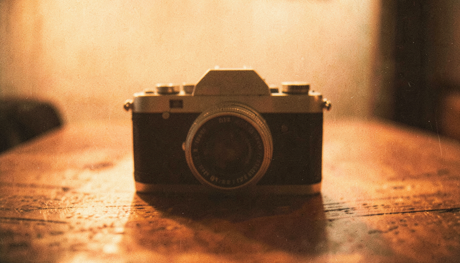 A warm-toned analog film photograph with visible grain, showing the soft silhouette of a vintage camera resting on a wooden surface in amber light.
