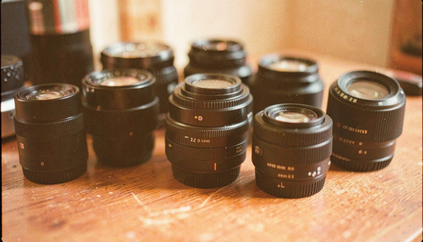 Warm-toned analog film close-up of assorted camera lenses arranged on a wooden surface, soft daylight, muted orange and brown tones