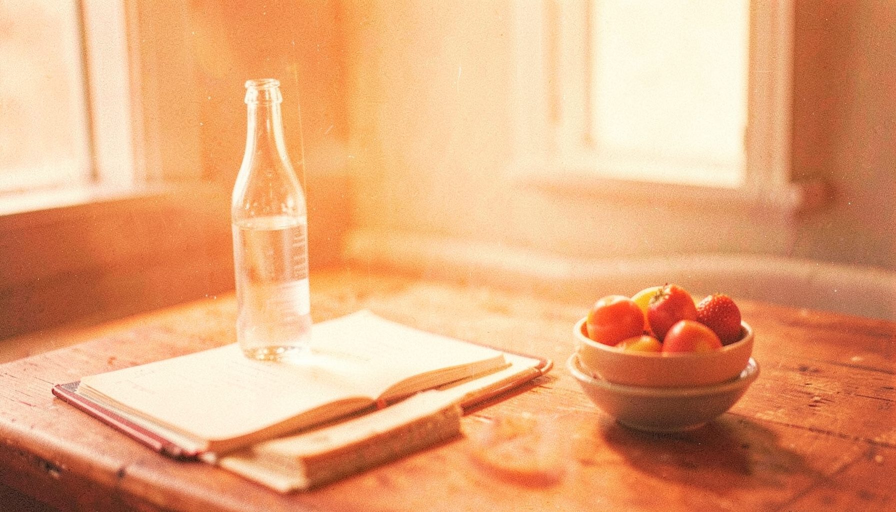 Warm orange and cream toned analog photograph of a still life with a glass water bottle, an open notebook, and a small book on a wooden table, soft natural light with visible film grain.