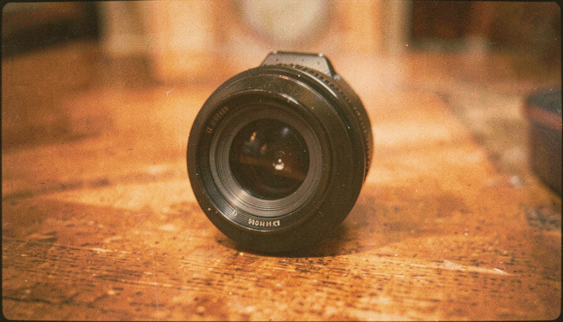 Analog film photograph of a camera lens resting on a wooden surface, warm amber and brown tones with visible grain